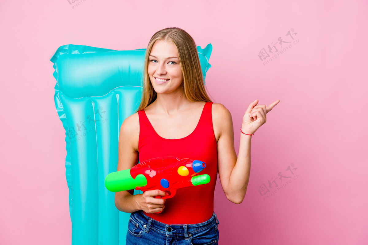 比基尼一个年轻的俄罗斯女人在玩一把水枪 手里拿着一个充气床垫 微笑着指着一边 在空白处展示了一些东西泳装身体手势