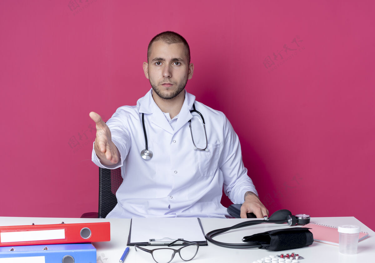 手势面容严肃的年轻男医生穿着医用长袍和听诊器坐在办公桌旁年轻坐着严肃