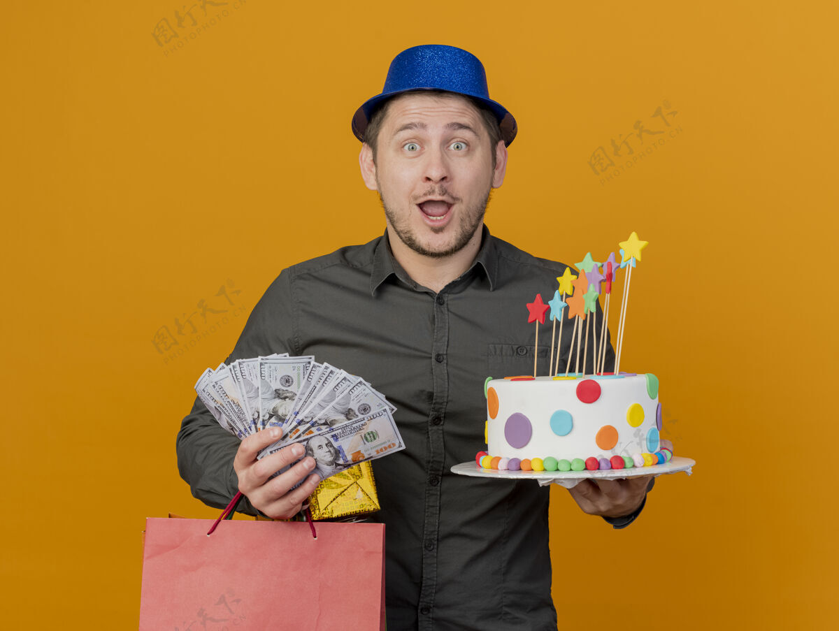 年轻人惊讶的年轻人戴着蓝色帽子拿着蛋糕和礼品袋 现金孤立在橙色穿上现金派对