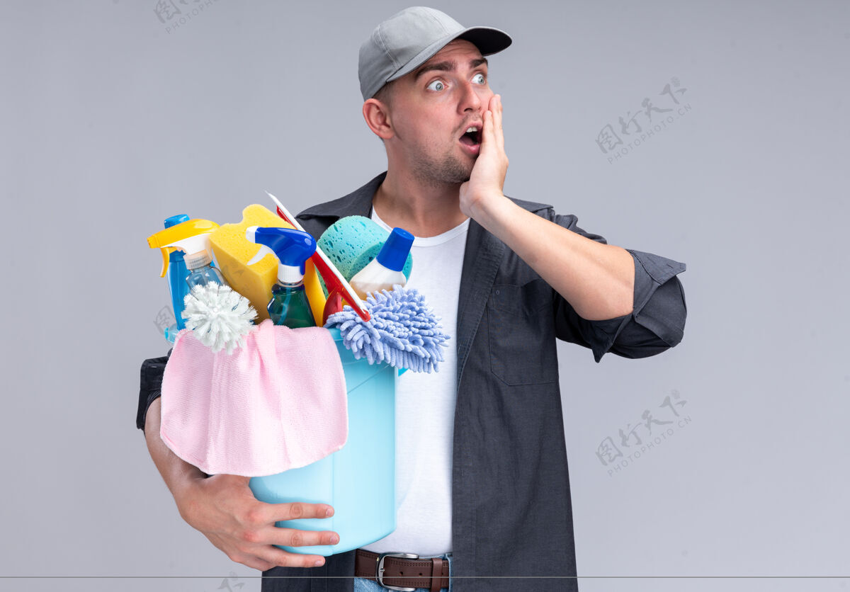 人年轻帅气的清洁工穿着t恤 戴着帽子 手里拿着一桶清洁工具 手放在隔离在白墙上的脸颊上脸颊衣服年轻