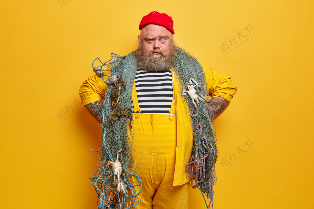 超重严肃的渔夫表情自信 双手放在臀部 穿着条纹水手衬衫和黄色工作服 准备用网捕鱼姿势职业人类