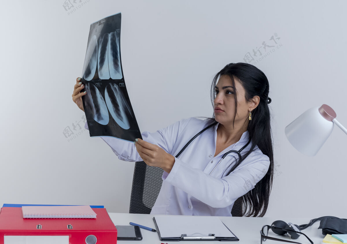 X光专注的年轻女医生穿着医用长袍和听诊器坐在办公桌旁 手持医疗工具 看着x光片工具坐着医疗人员