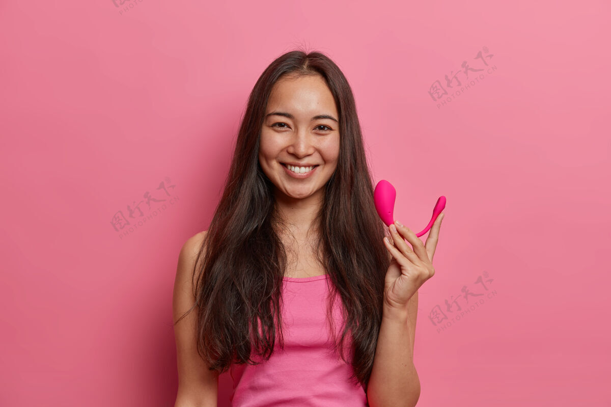 情绪快乐的黑发女人用智能蓝牙振动器摆出姿势 用手机上的特殊应用程序来提高性高潮 拿着性爱工具来增强快感 隔离在粉红色的墙上现代科技和性生活高兴性现代