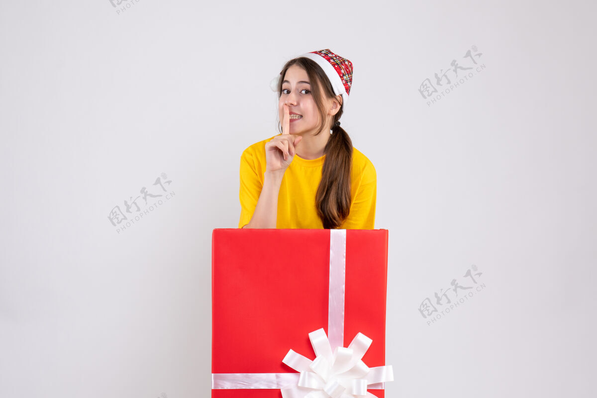 站前视图可爱的女孩与圣诞帽制作嘘标志站在大圣诞礼物后面圣诞节微笑丝带