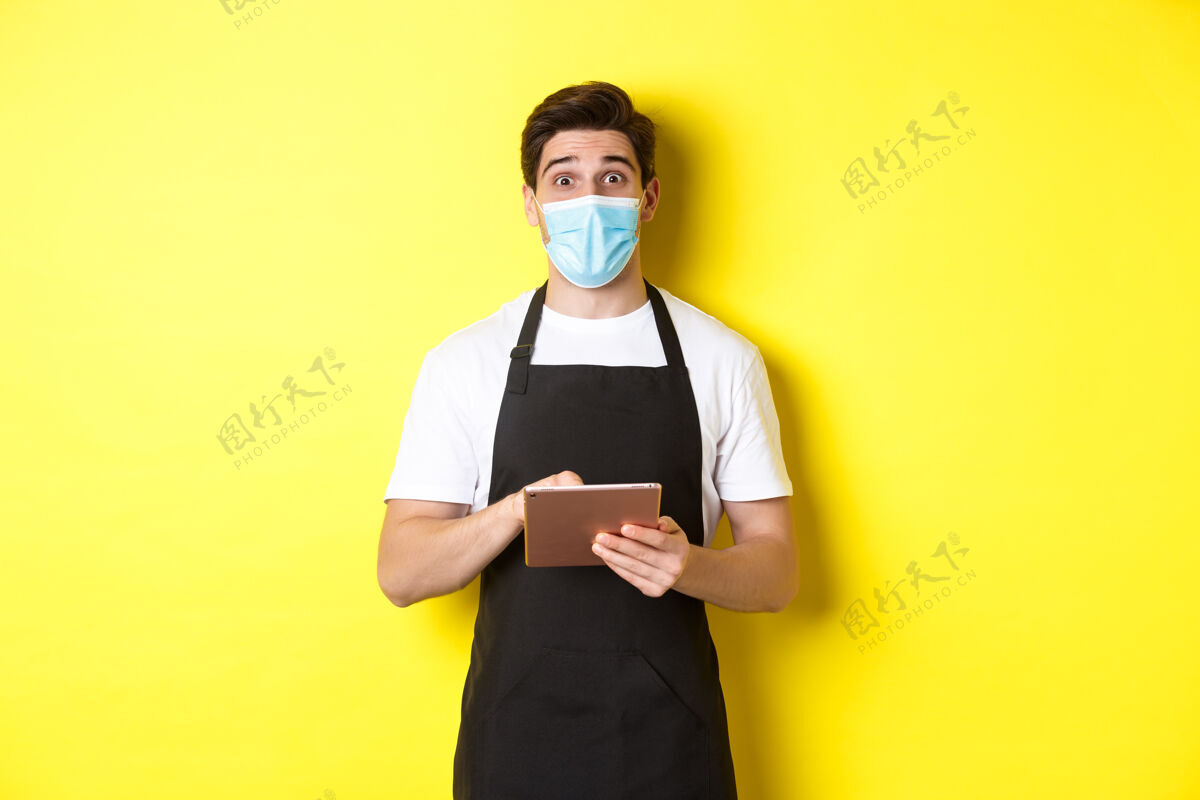 平板电脑covid-19的概念 小型企业和流行病穿着黑色围裙和医用面罩的服务员拿着数字平板电脑 站在黄色背景上帅哥男士男士