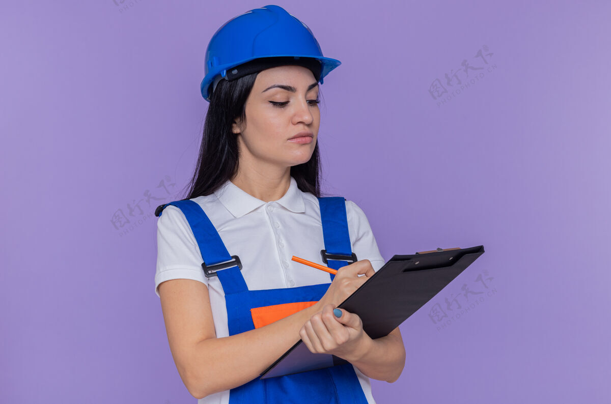 拿着身穿建筑制服 头戴安全帽的年轻建筑工人站在紫色的墙上 手里拿着剪贴板 写着一些看起来很自信的东西写作建筑女人