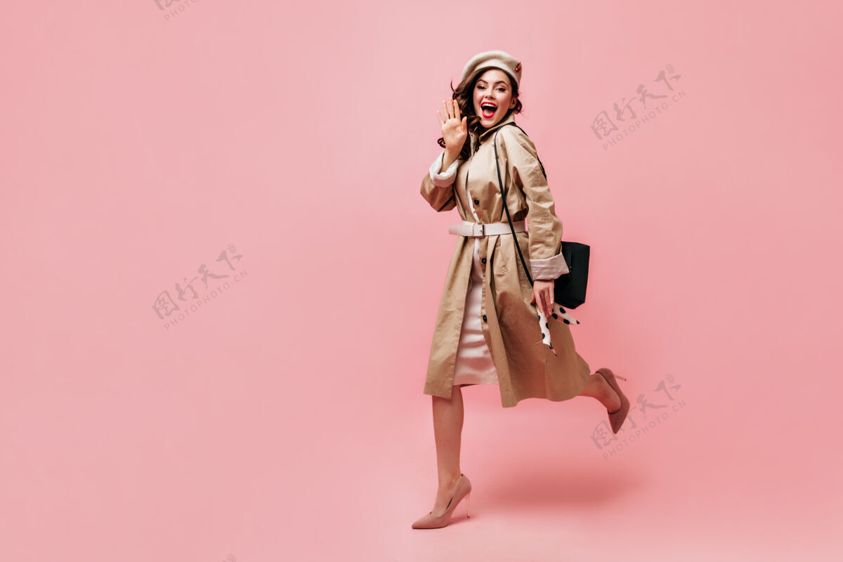 年轻穿着风衣和贝雷帽的女孩高兴地挥舞着她的手 在粉红色的背景上奔跑优雅波浪发海沟