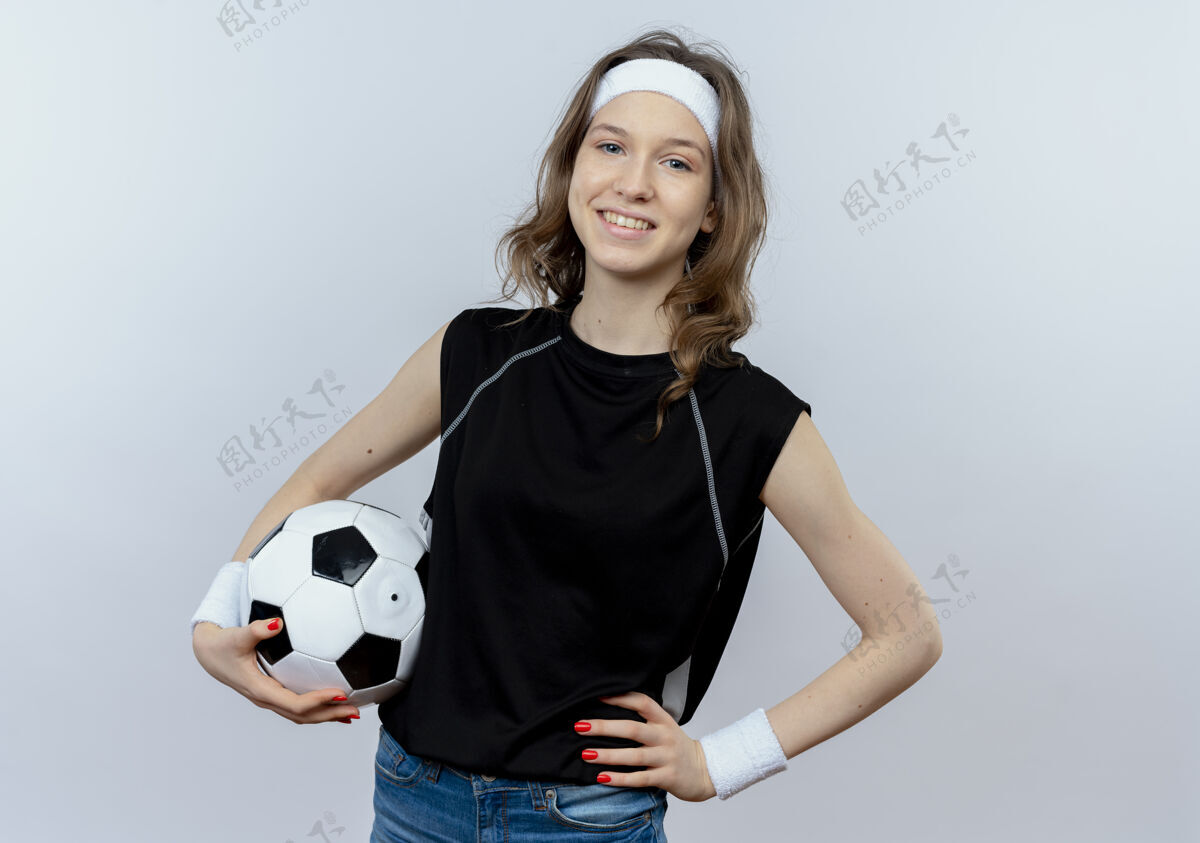 球身穿黑色运动服 头箍 手持足球 自信地站在白墙上的年轻健身女孩健身微笑女孩