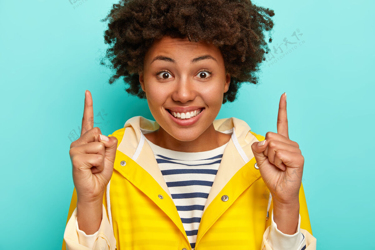 黄色笑容满面的非裔美国妇女 表情愉快 向上显示方向 穿着条纹套头衫和黄色雨衣 隔离在蓝色背景下蓝色满意食指