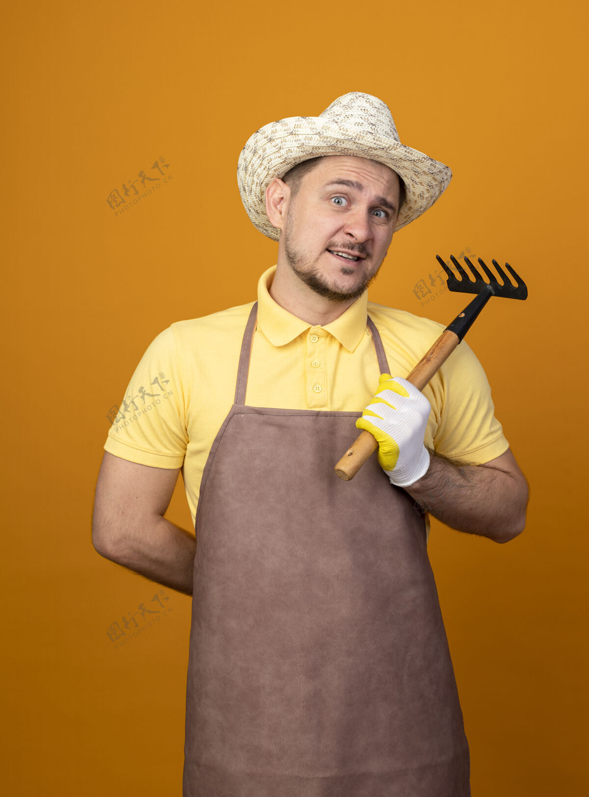 困惑年轻的园丁穿着连体衣 戴着帽子 手里拿着小耙子 站在橙色的墙上 困惑地微笑着看着前面市民工人连身衣