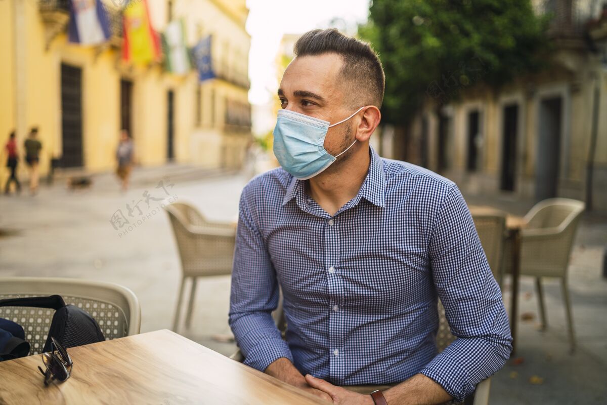 外科身穿蓝色衬衫 戴着医用面罩的年轻男子坐在室外咖啡馆里现代智能健康