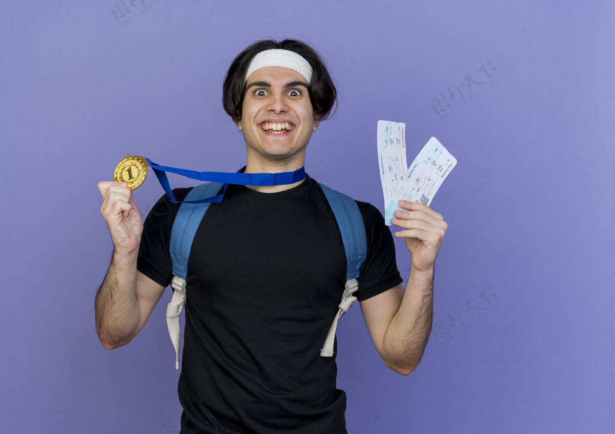 穿着一个年轻的运动型男人 穿着运动服 戴着头巾 背着背包 脖子上挂着金牌 手里拿着机票 看着镜头 笑得发疯 开心运动装头带背包