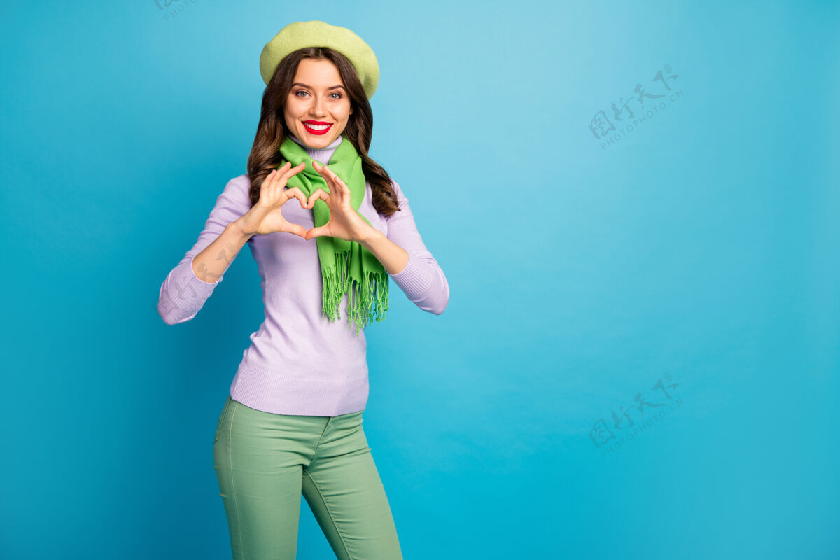 天照片中搞笑迷人的女士手牵手心形象征心意穿着时尚的绿色贝雷帽紫色高领围巾长裤孤立的蓝色墙壁情人喜欢休闲