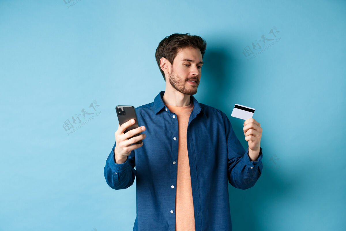 男性电子商务概念男人在线支付 读信用卡号 拿着手机 站在蓝色的背景上胡须发型移动