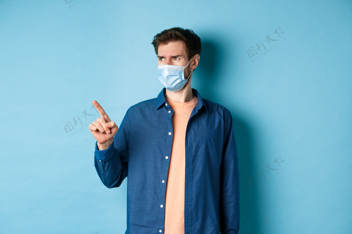 面具Covid-19和医疗保健概念困惑和怀疑的人戴着面具皱着眉头 指着空荡荡的空间 站在蓝色的背景上流行病冠状病毒男性