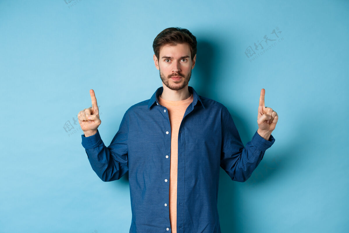年轻严肃的白种人 留着胡子 手指指着空旷的空间 展示标志 看着摄像机 站在蓝色的背景上手发型胡须