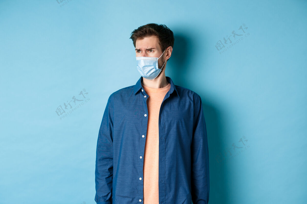 年轻Covid-19和医疗保健概念不满意皱眉的家伙戴着医用口罩看着不好的宣传片 带着失望的脸 蓝色的背景盯着空荡荡的空间英俊冠状病毒流行病
