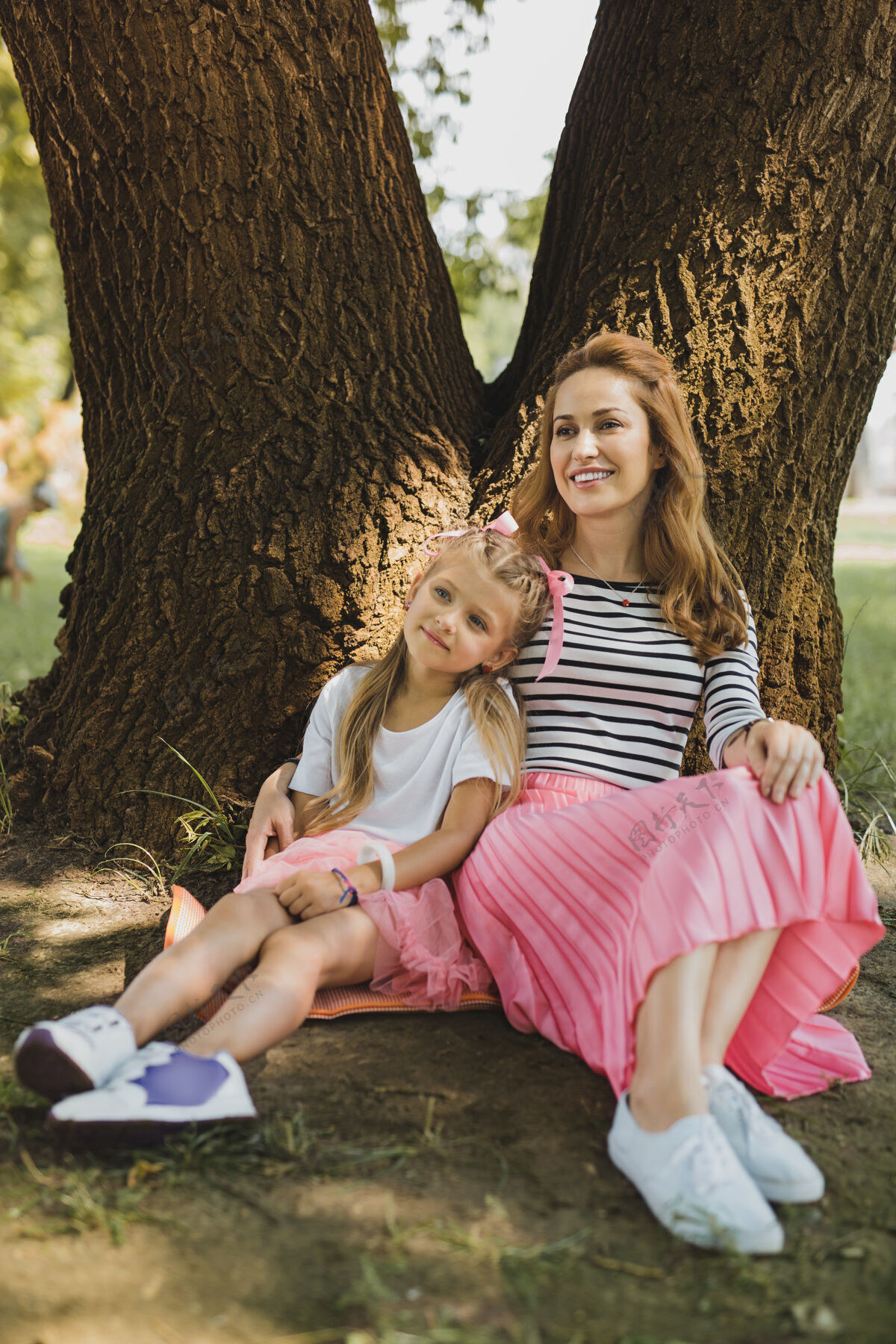 收养粉红色的裙子微笑的爱的女人穿着粉红色的裙子坐在她时髦的小女儿旁边一起坐着树
