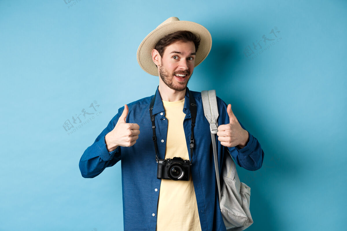 认可快乐微笑的游客说是的 竖起大拇指 去放暑假 拿着背包和照相机 蓝色的背景喜欢男成人