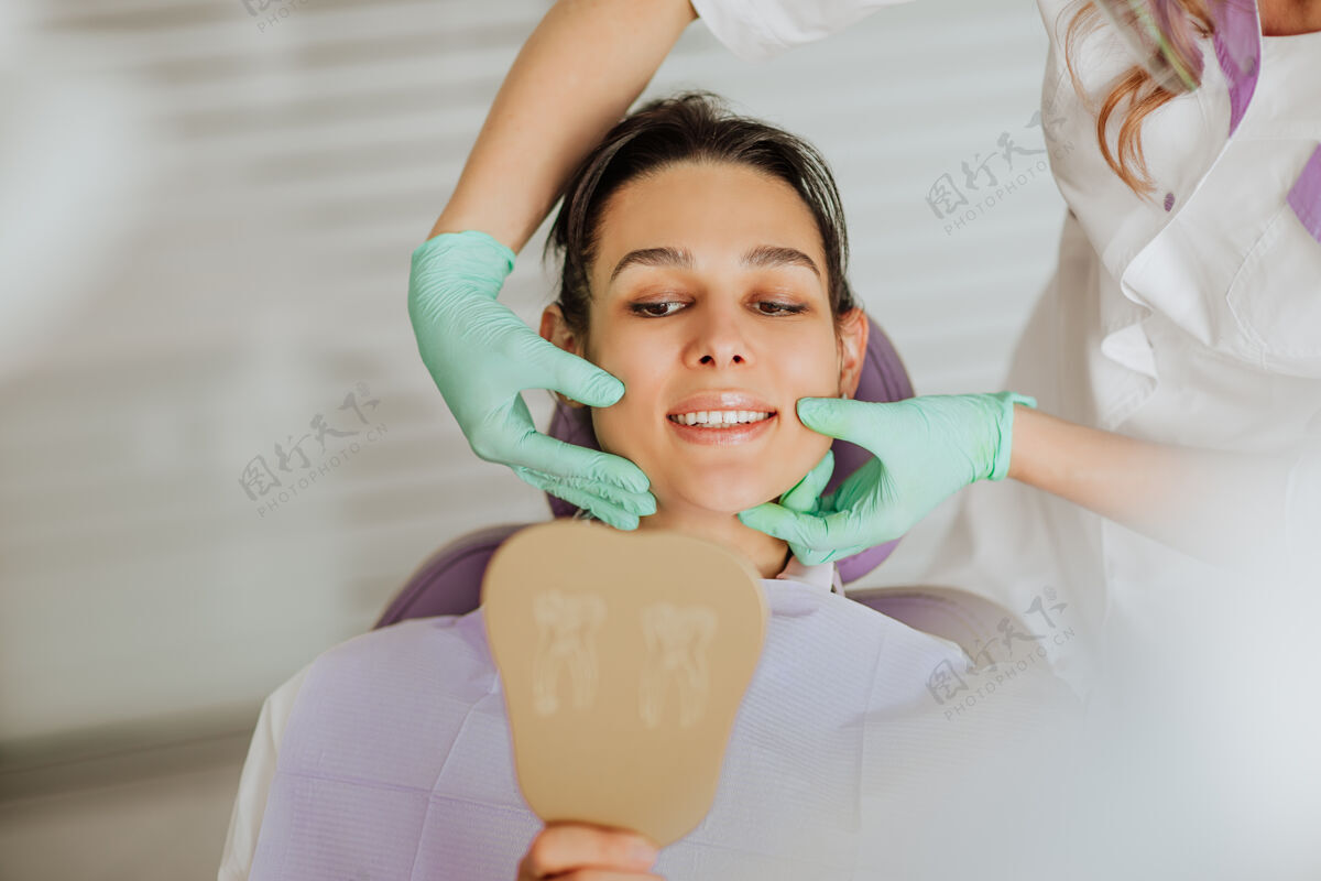 年轻人金发迷人的牙医医生戴着医用口罩和手套 与拿着镜子微笑的黑发女患者合影专业人士女士正畸