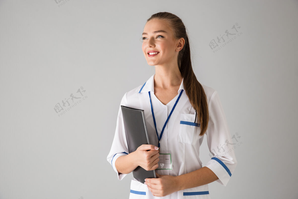 制服一位美丽快乐的年轻女性美容师医生的照片被隔离在灰色的墙上拿着文件夹工人专家医学