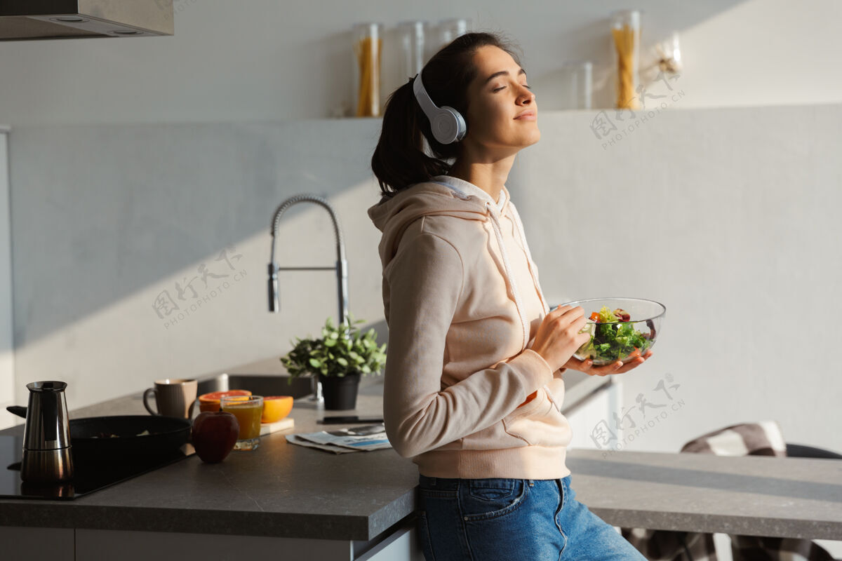 休闲快乐健康的女人每天早上站在厨房吃沙拉 戴着耳机听音乐早餐快乐肖像