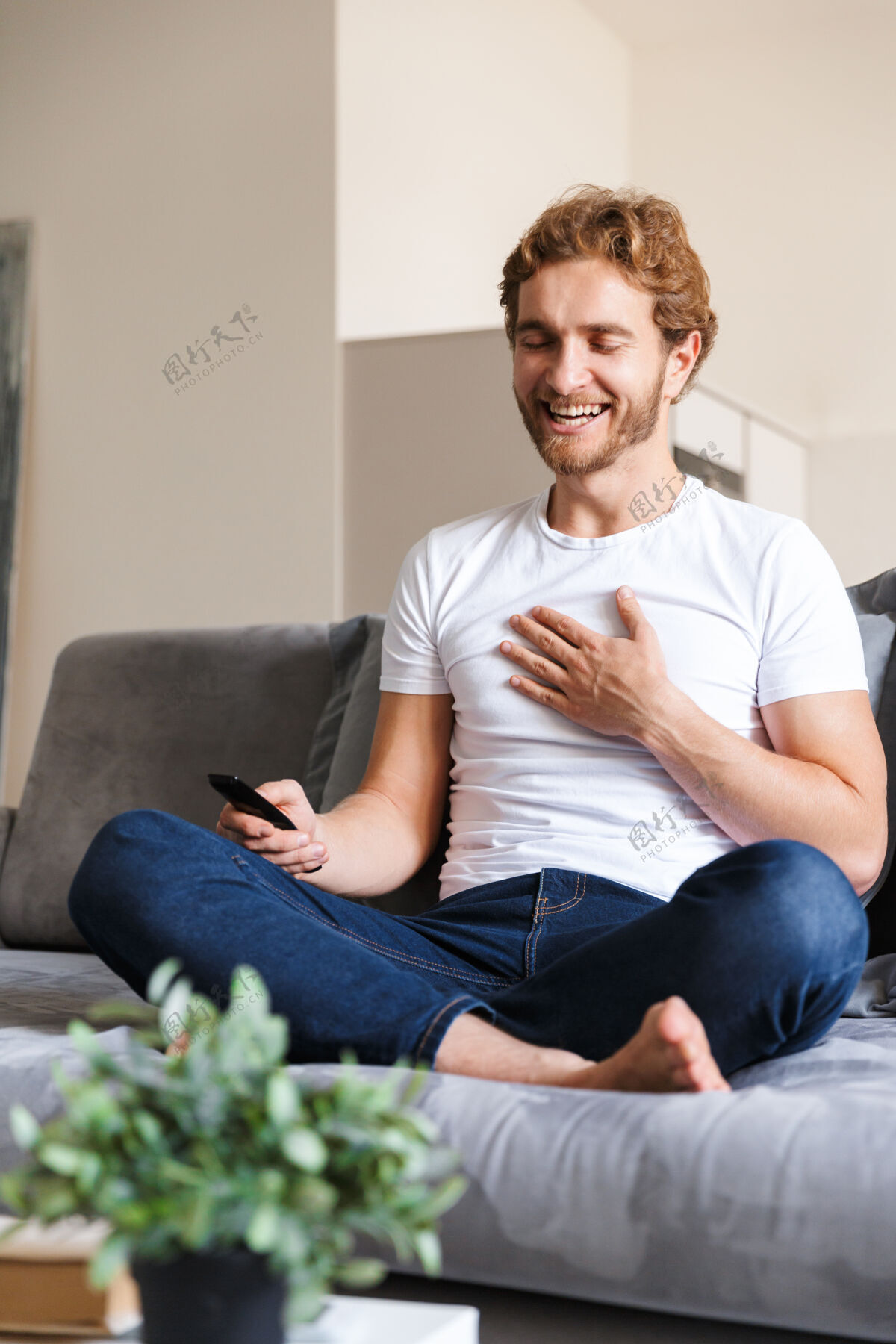 舒适一个英俊快乐的年轻人在家里的沙发上拿着遥控器看电视软文学沙发