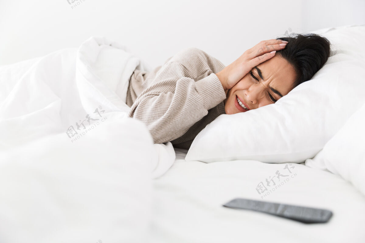 抓取一张30多岁的女人因为智能手机而抓头的照片 她躺在床上 家里穿着白色亚麻布室内电话消遣