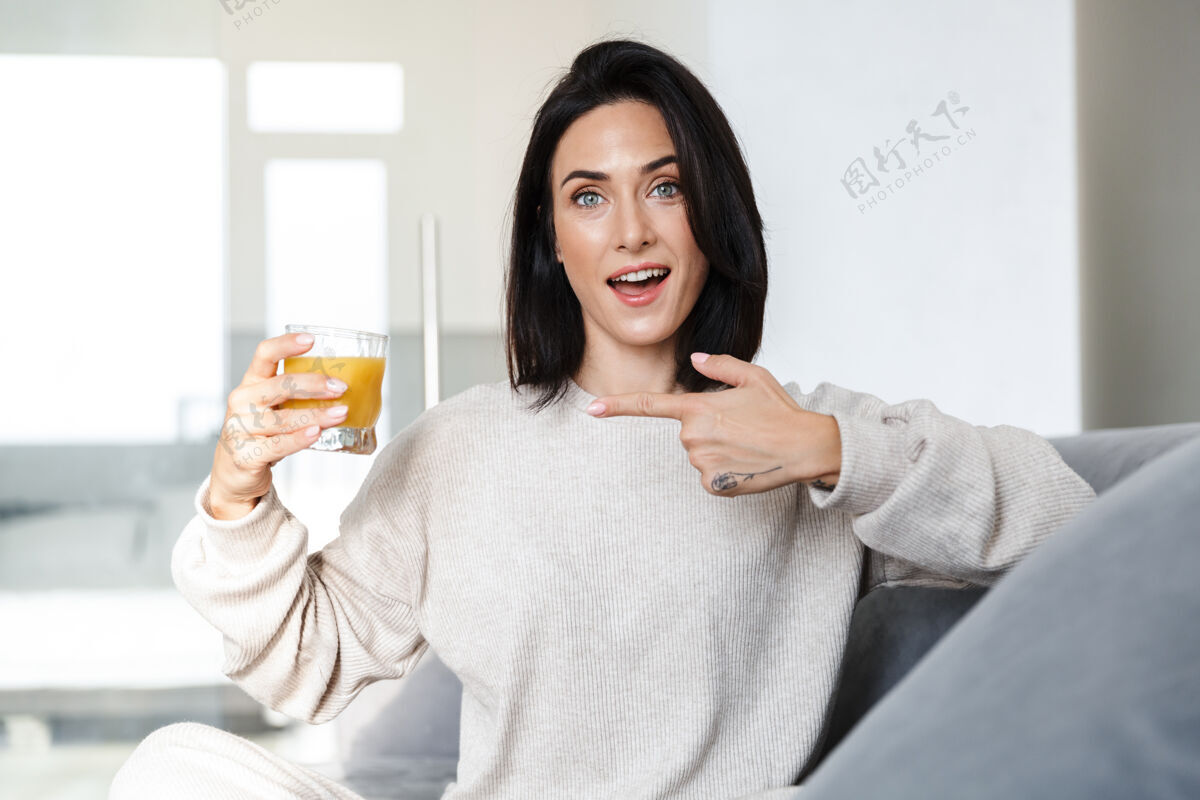 舒适一张30多岁的欧洲女人在明亮的房间里坐在沙发上喝橙汁的照片玻璃愉快饮用