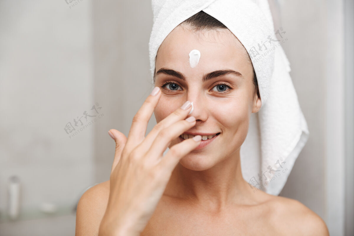 化妆特写镜头：一个头上戴着毛巾的年轻漂亮女人站在浴室里涂面霜微笑浴室女人