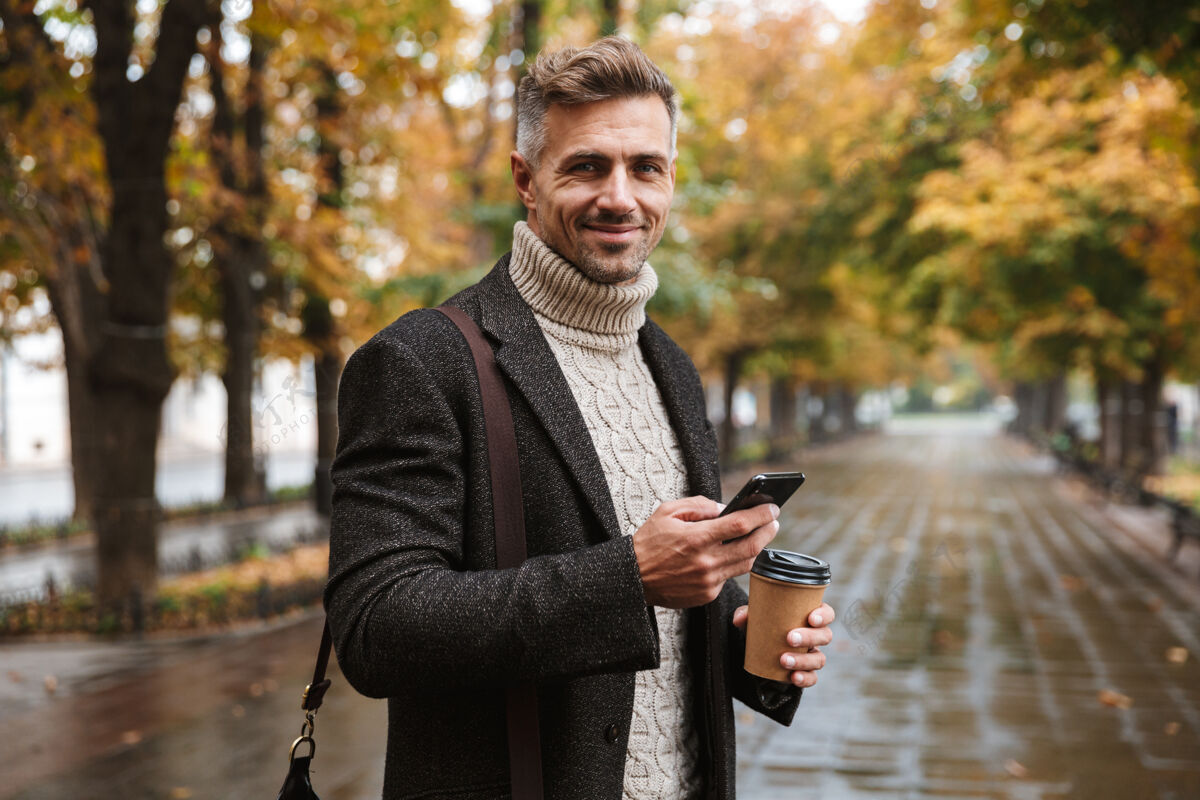 寒冷30多岁的帅哥穿着暖和的衣服在户外穿过秋园 用手机拍照公园男士享受