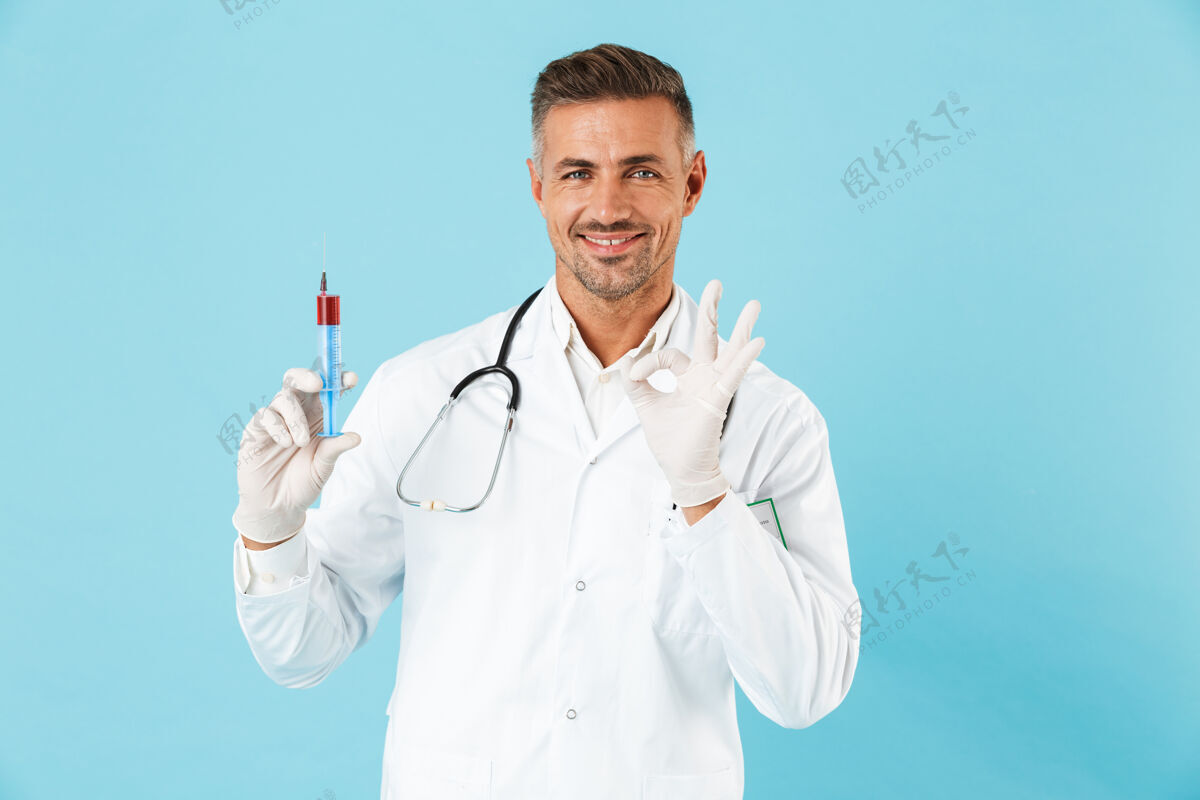 实验室自信的男医生穿着制服 孤立地站在蓝色的墙上 展示着一个装有红色液体的注射器职业成人制服