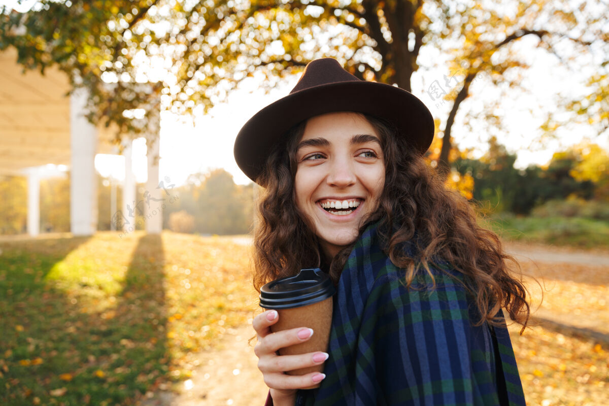 温暖美丽的年轻女子戴着帽子在秋天的公园散步 手里拿着一杯外卖咖啡公园户外享受