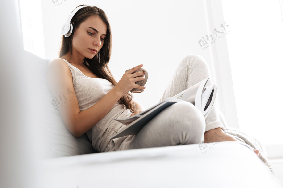 阅读美丽的年轻女子坐在家里的沙发上 戴着耳机听音乐 喝茶 看杂志地毯休息马克杯