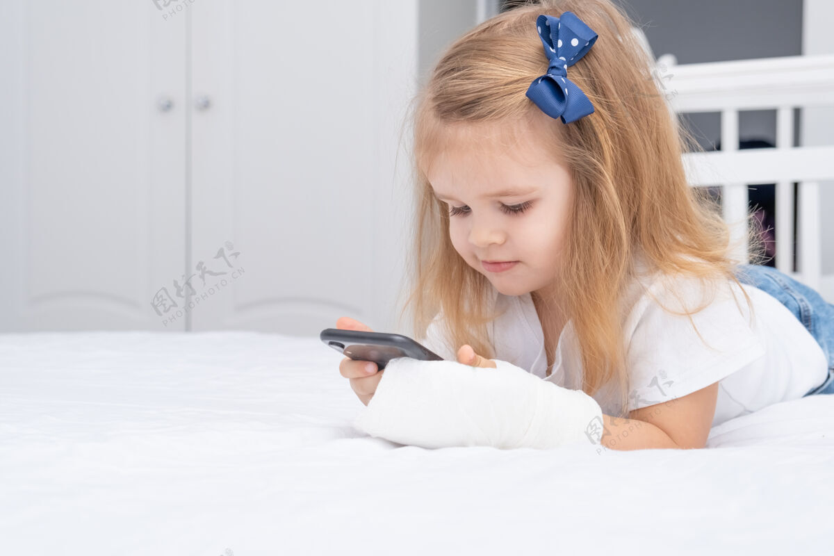 卡通手拿石膏的小女孩躺在床上用智能手机 看动画片或教育视频治疗事故石膏