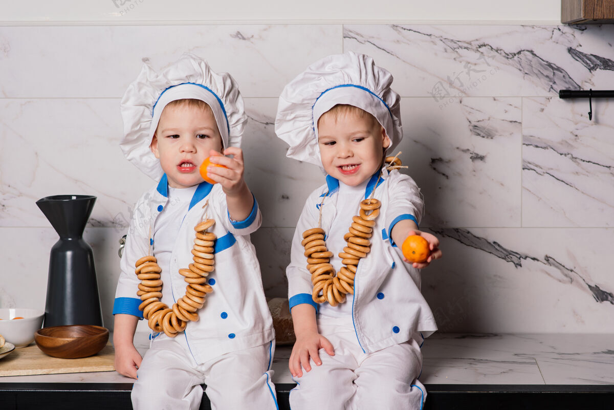 青少年小可爱的厨师拿着餐具坐在厨房里 双胞胎兄弟 橘子 百吉饼有趣糕点庆祝