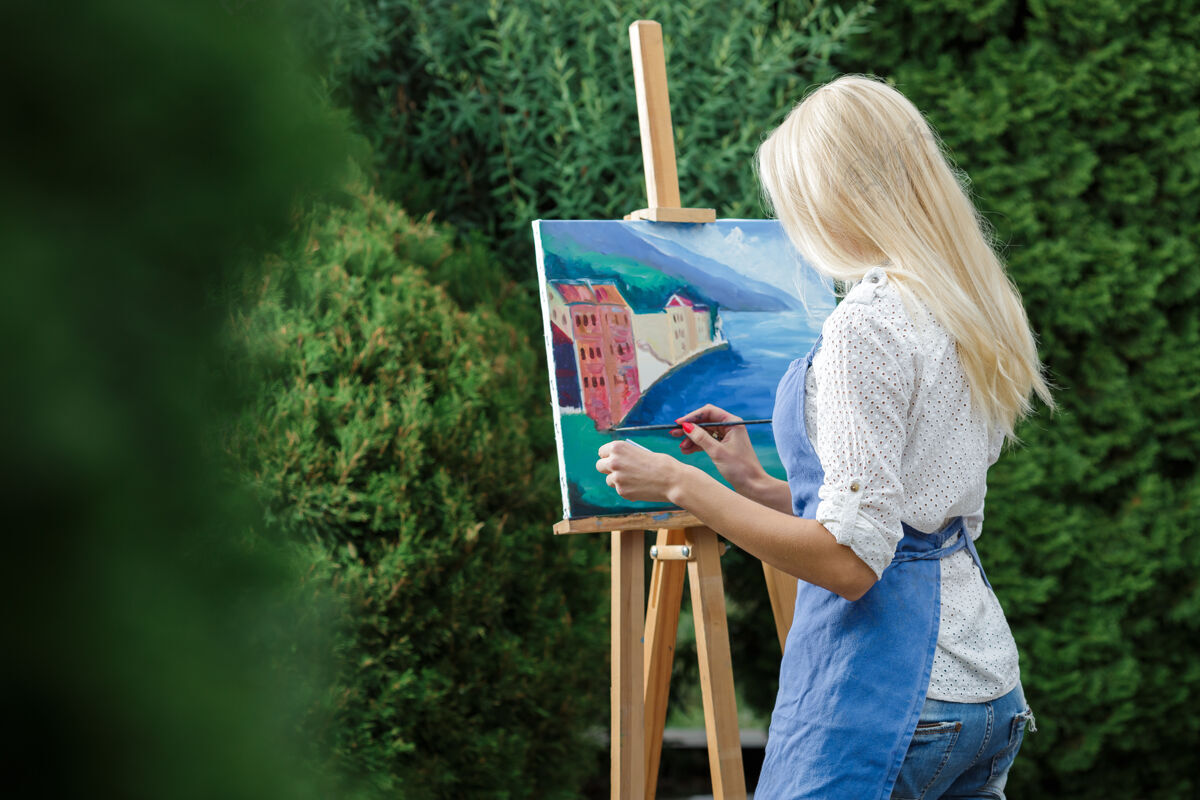 画架美丽的金发女画家手里拿着画笔在花园里的画布上画画色彩背景灵感