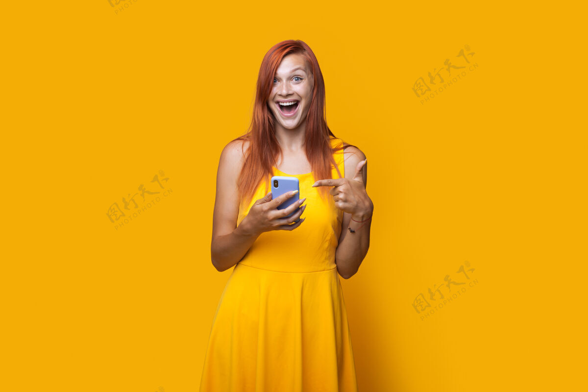 姜黄色工作室墙上 一位穿着裙子 红头发的白人妇女指着手机的单色照片技术生活方式电话