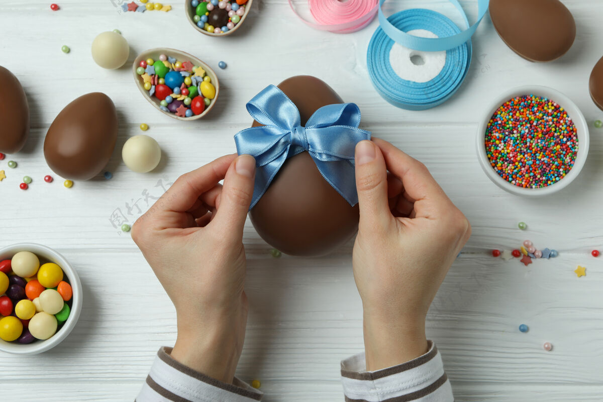 装饰女人的手拿着蝴蝶结在白色木制的巧克力蛋上 上面有复活节的饰物庆典糖果复活节