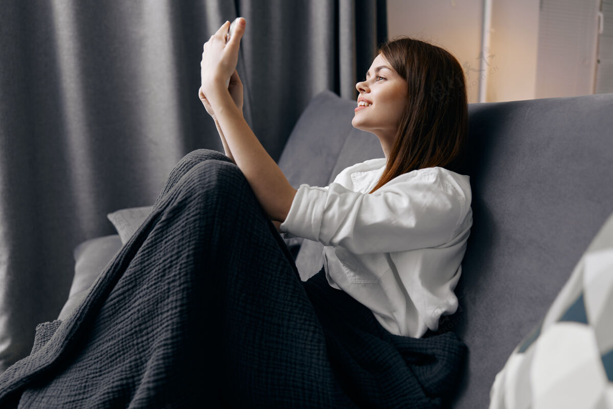 电话快乐的女人看着手机屏幕 坐在室内的沙发上房子便携式博客