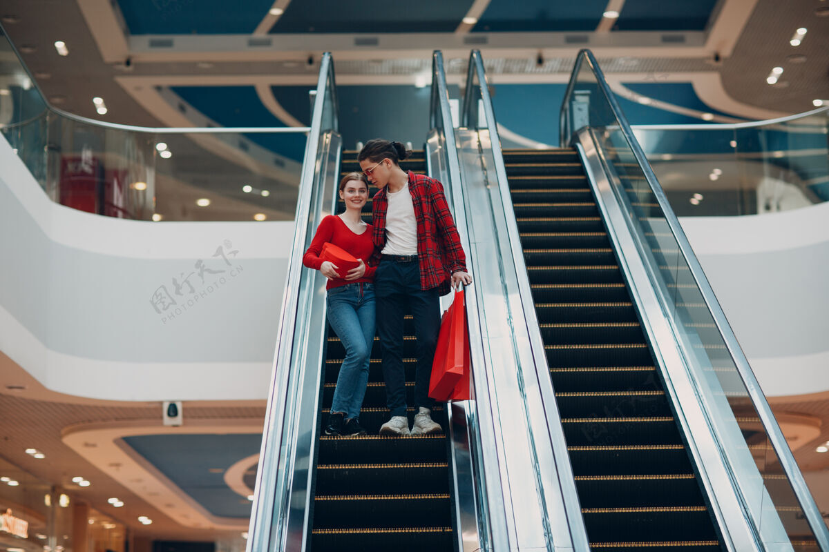 女人年轻美丽幸福的年轻夫妇 带着购物袋在商场的自动扶梯上中心精品店情侣