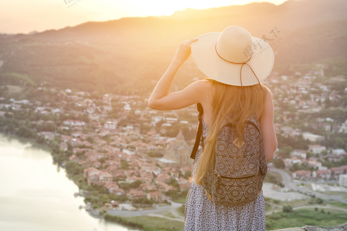 女人一个背着背包 戴着帽子的女人看着下面的城市和河流背景帽子日落