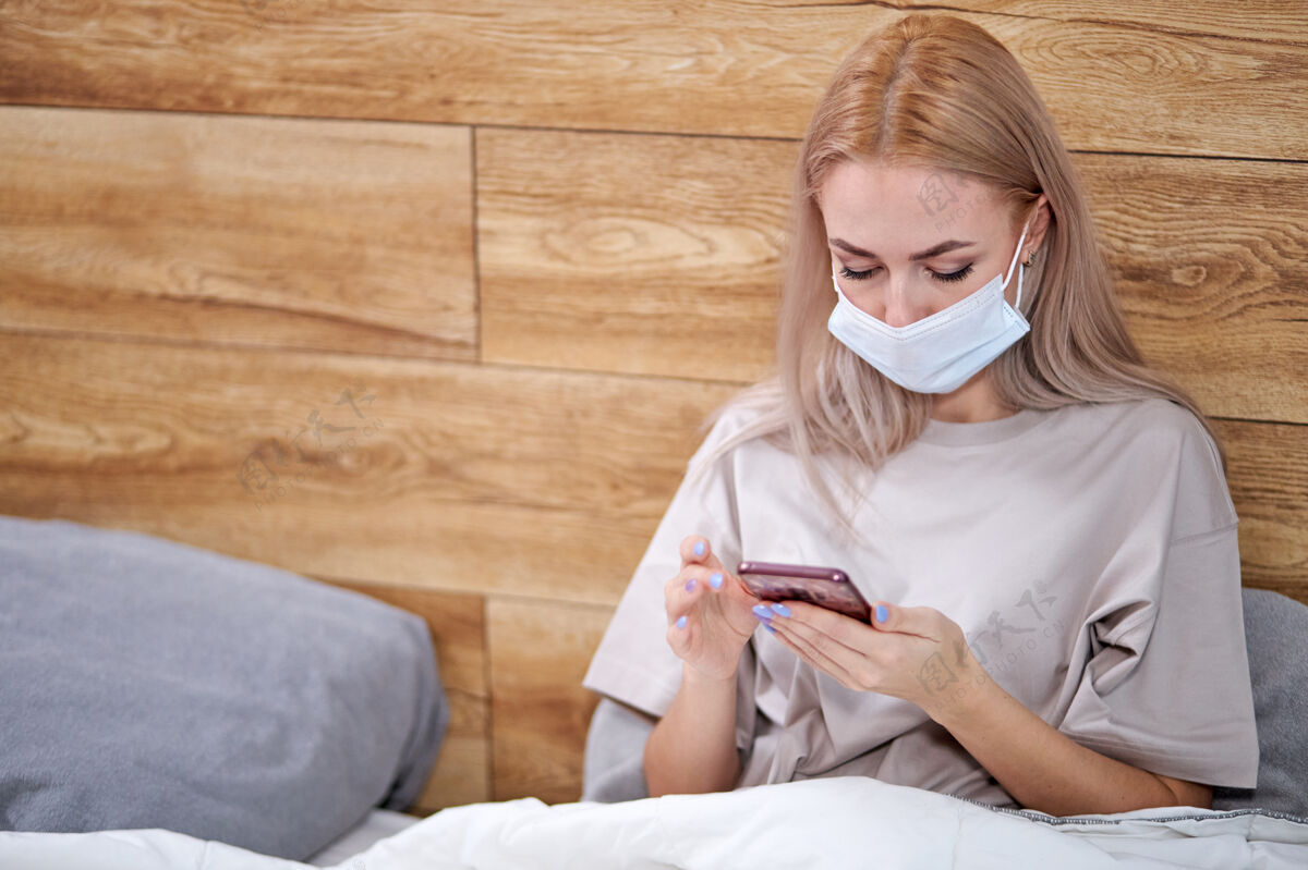 电话戴着医用口罩的妇女躺在床上 带着智能手机 在自己被隔离时休息-冷隔离流感 发烧和偏头痛年轻护理坏