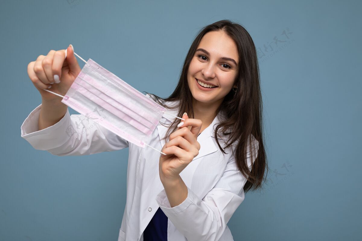 女性一位面带微笑的女士 手拿防冠状病毒的防毒面具 身穿蓝色背景隔离的白色医用外套医生大流行白大衣