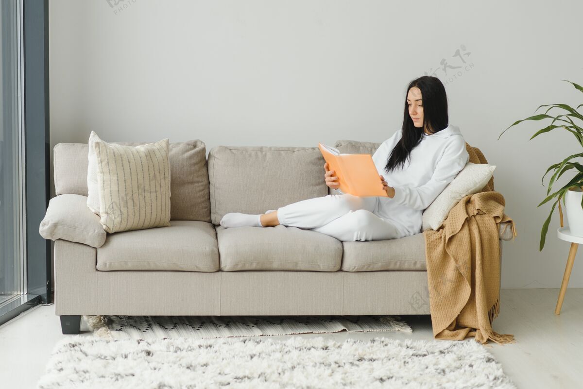 室内漂亮的年轻女子躺在沙发上享受着在家看书的乐趣 穿着休闲装微笑着坐着室内肖像