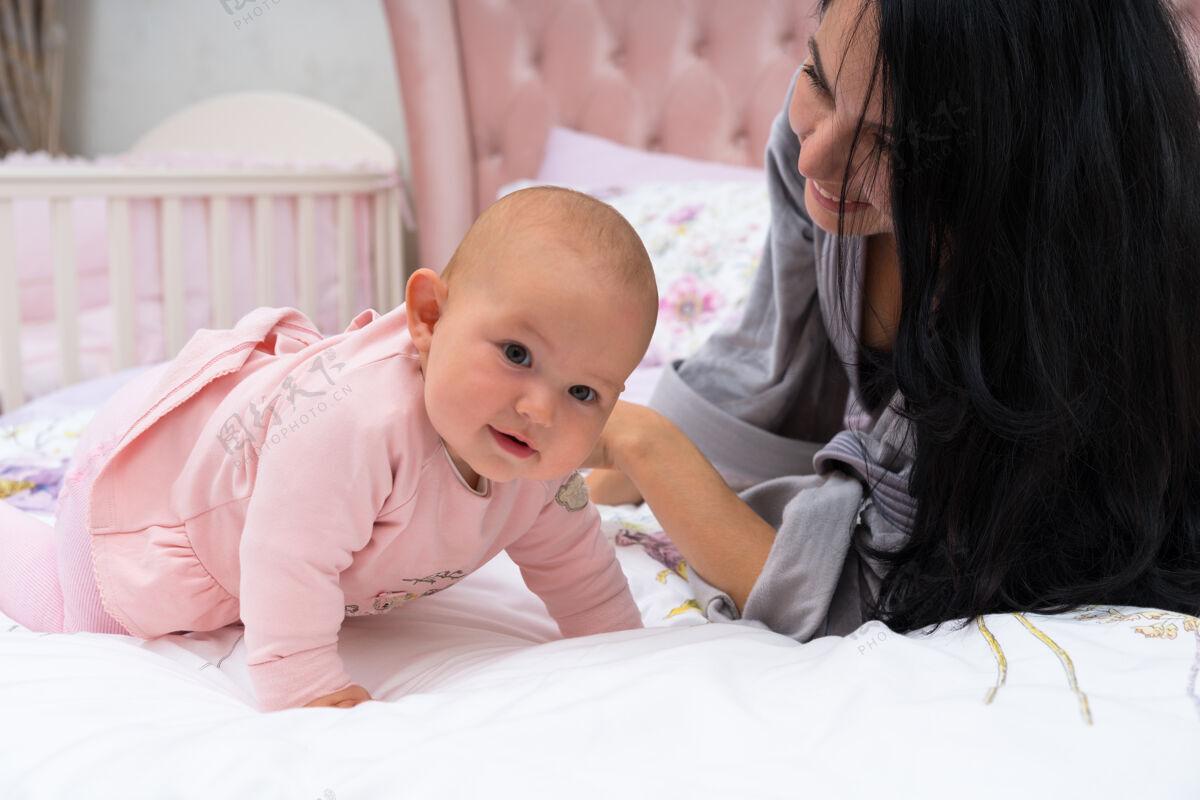 站着穿着浴衣的年轻妈妈躺在床上 和穿着粉色连衣裙的可爱快乐的女婴一起享受时光 面带微笑婴儿卧室情绪