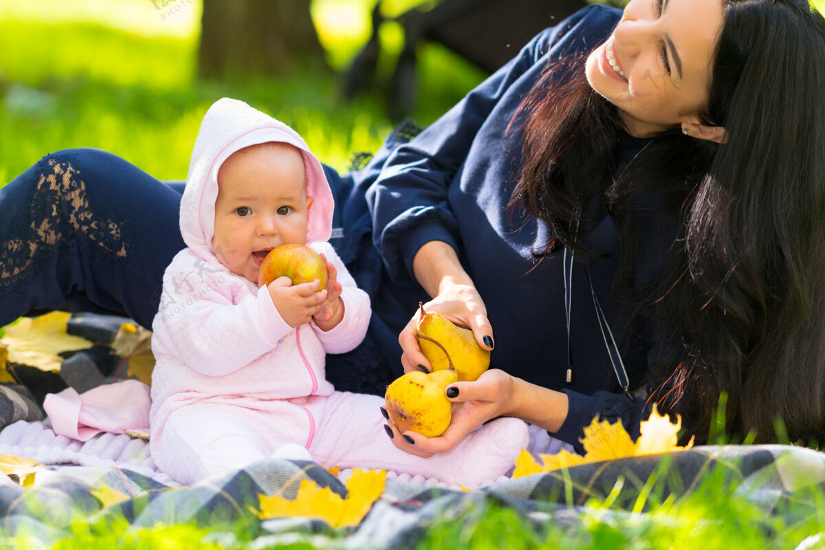 刺激在一个秋天的公园里 年轻快乐的妈妈抱着一个成熟的金苹果 坐在草地上的地毯上 笑着她的小女儿母性女孩邦德