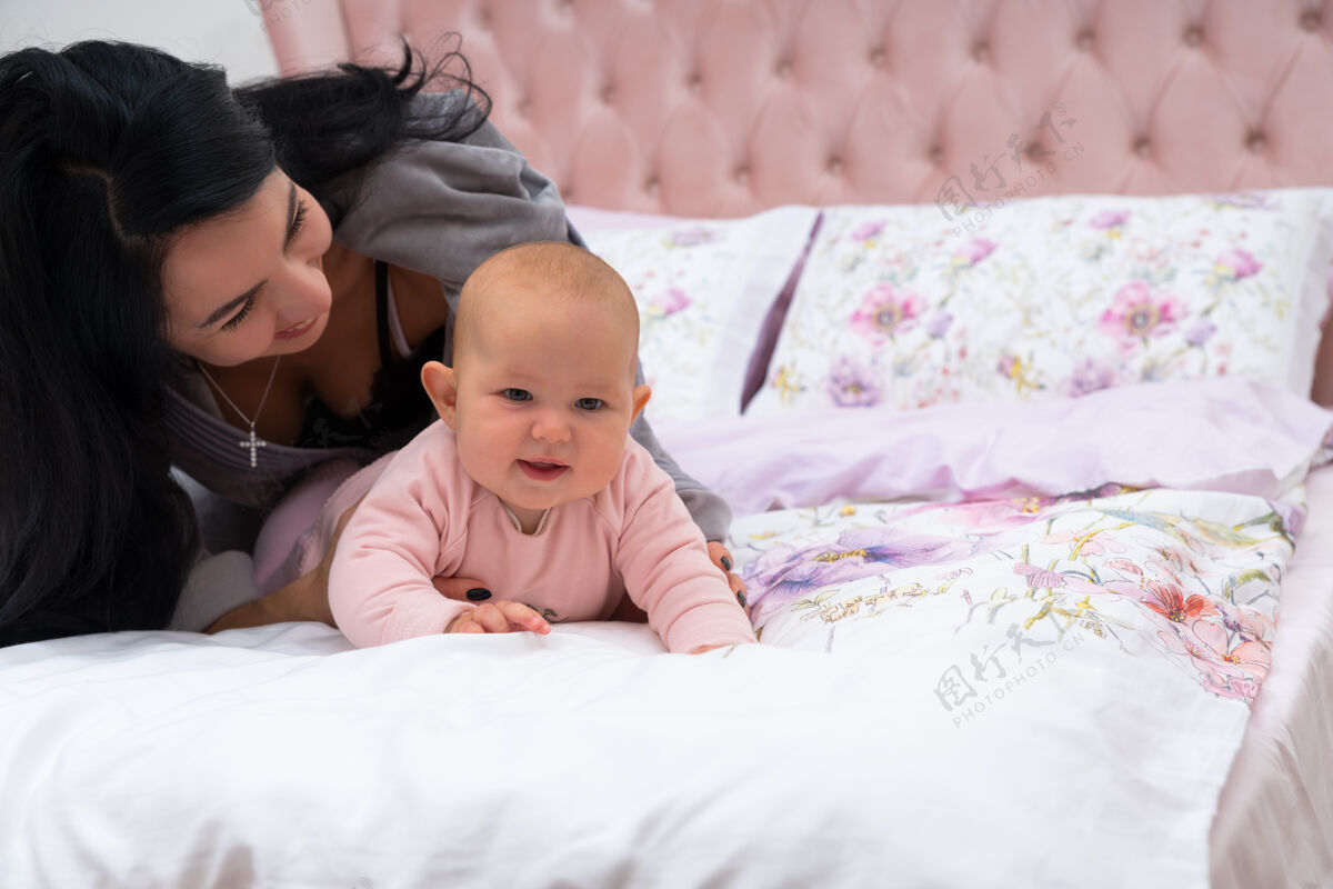 室内妈妈在床上玩她的孩子 而小女婴正试图爬着离开她的微笑面对面正面拍摄 复制空间抱着快乐在一起