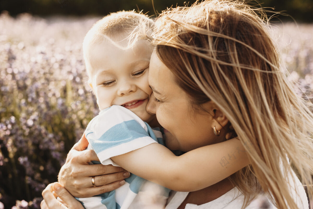 关系一位年轻母亲紧抱着儿子的特写照片 她闭着眼睛 微笑着站在户外的花丛中 夕阳映照女性乐趣肖像