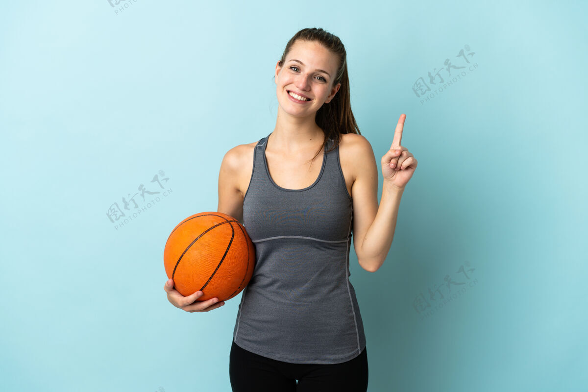 手势在蓝色背景上孤立地打篮球的年轻女子展示并举起手指表示最棒爱好年轻手